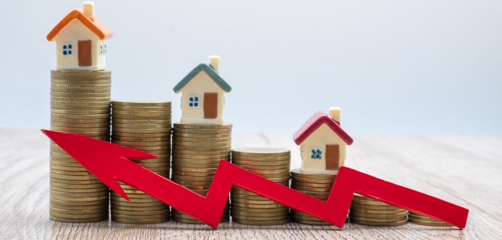 Stacks of coins with small house models on top and a red downward arrow in front, echoing the findings from the Manitoba Quarterly Market Overview that highlights a decline in the housing market.