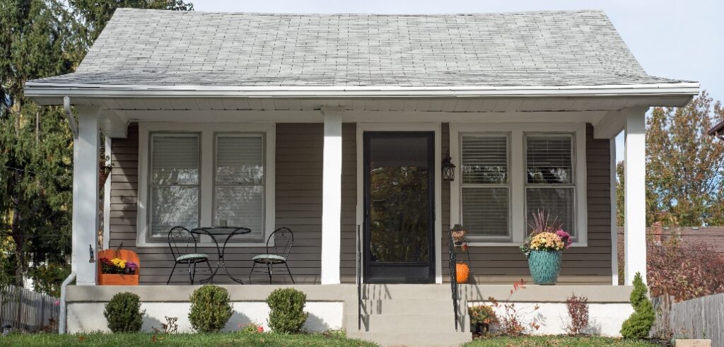 A small, single-story house with a gray roof and beige siding, showcasing the charm of new housing construction. A front porch features two black metal chairs, a table, and potted plants. There are shrubs and a well-maintained lawn in the front yard.