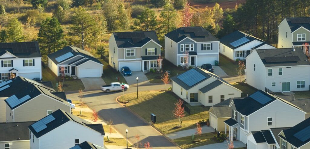 Aerial view of a suburban neighborhood showcasing housing with multiple two-story houses, some with solar panels. Cars are parked in driveways, and trees are scattered throughout the area.