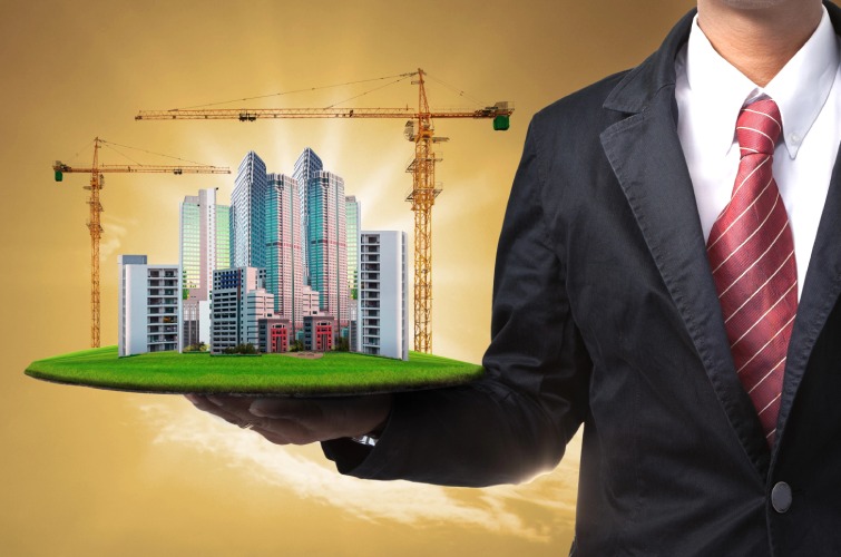A person in a suit holds a model of a city with high-rise buildings and construction cranes on a flat surface.