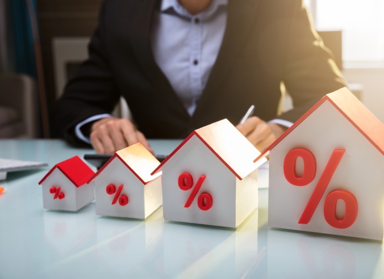 A person in a suit is meticulously writing on paper at a desk, with four different-sized house models displaying percentage symbols in the foreground, illustrating mortgage rates.