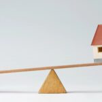 A model house and stacks of coins are balanced on opposite ends of a wooden seesaw, illustrating the concept of financial balance in real estate investments.