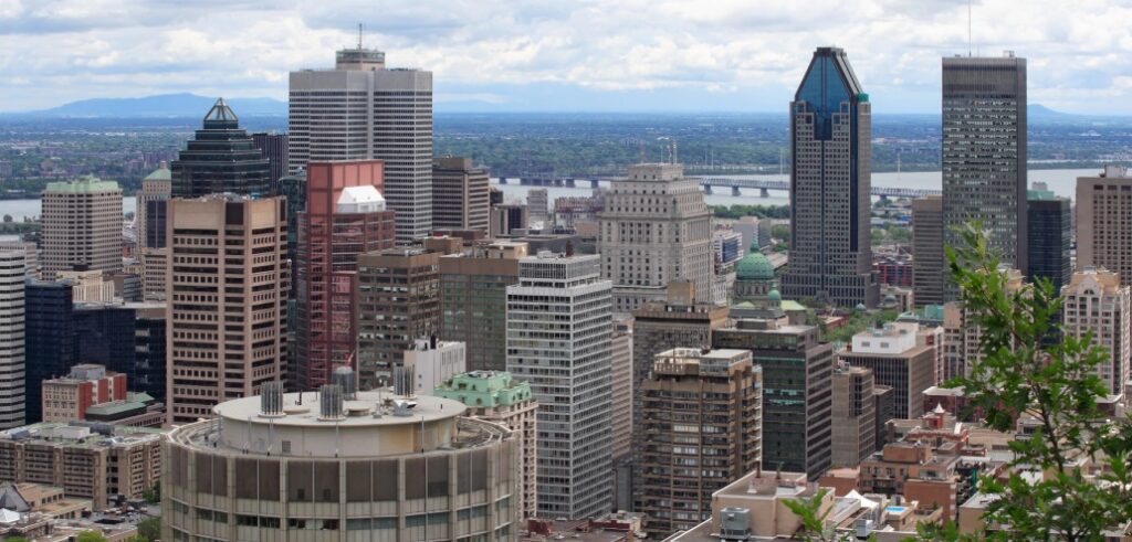 Aerial view of downtown Montreal Metropolitan Area featuring various high-rise buildings with mountains and the St. Lawrence River visible in the background under a partly cloudy sky.