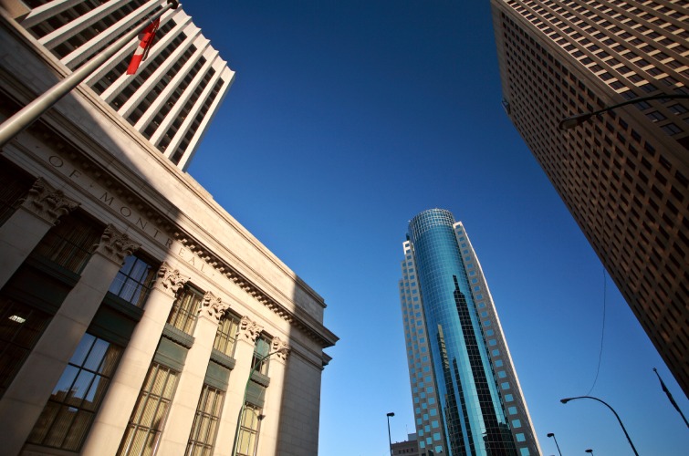 View of four tall buildings against a clear blue sky, looking upwards from street level. One building has a flag on a pole in the foreground.