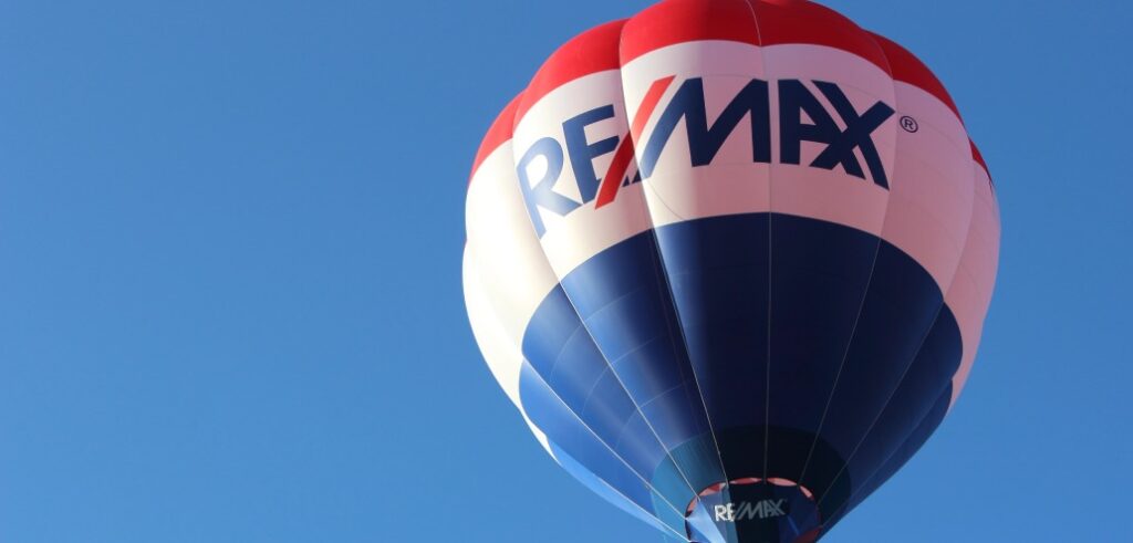 A red, white, and blue hot air balloon with the RE/MAX logo floats against a clear blue sky.