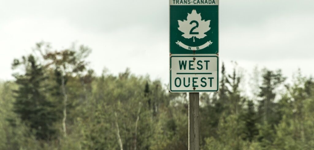 A road sign on a rural highway shows "Trans-Canada 2, NB, West/Ouest" with trees and a cloudy sky in the background.