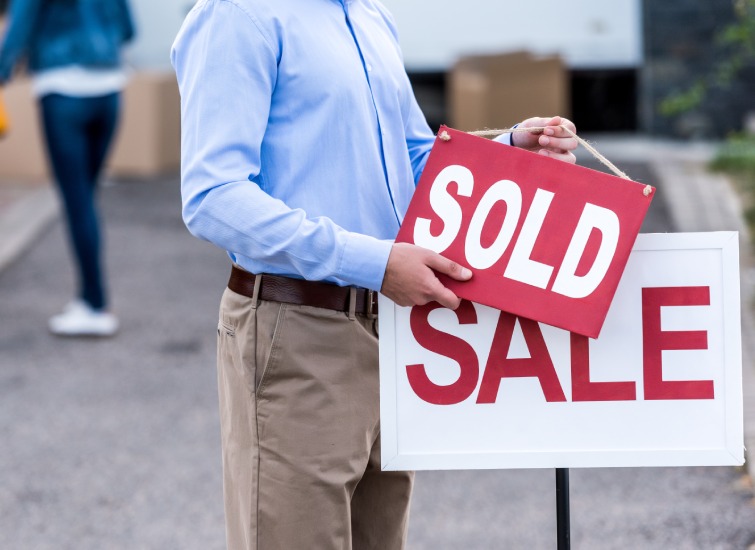A person in a blue shirt is holding a red "SOLD" sign over a "SALE" sign outdoors in Saskatchewan, with someone else blurred in the background.