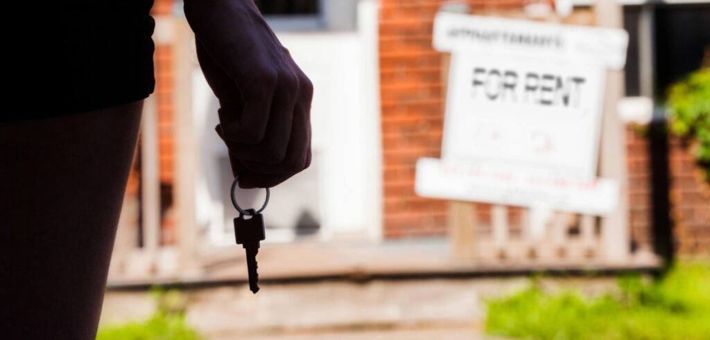 Person holding a key in front of a house with a "For Rent" sign.
